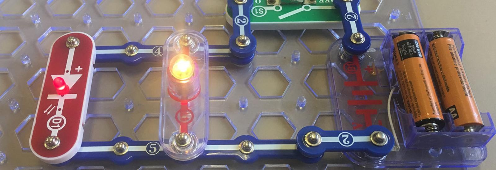 Photograph of a closed parallel circuit created using snap circuits, an LED diode, light bulb, switch, and batteries. 