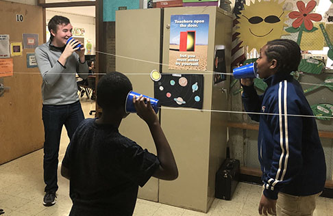 Photograph of a volunteer teaching 5th graders about sound using cups to play telephone