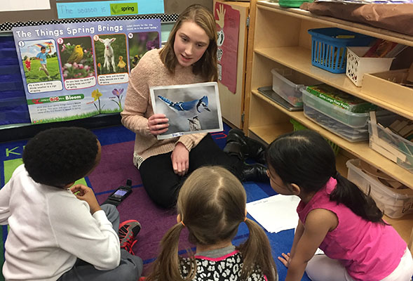 Photograph of undergrad volunteer teaching preschoolers about bird songs. The volunteer is also holding a photo of the bird, a Blue Jay.